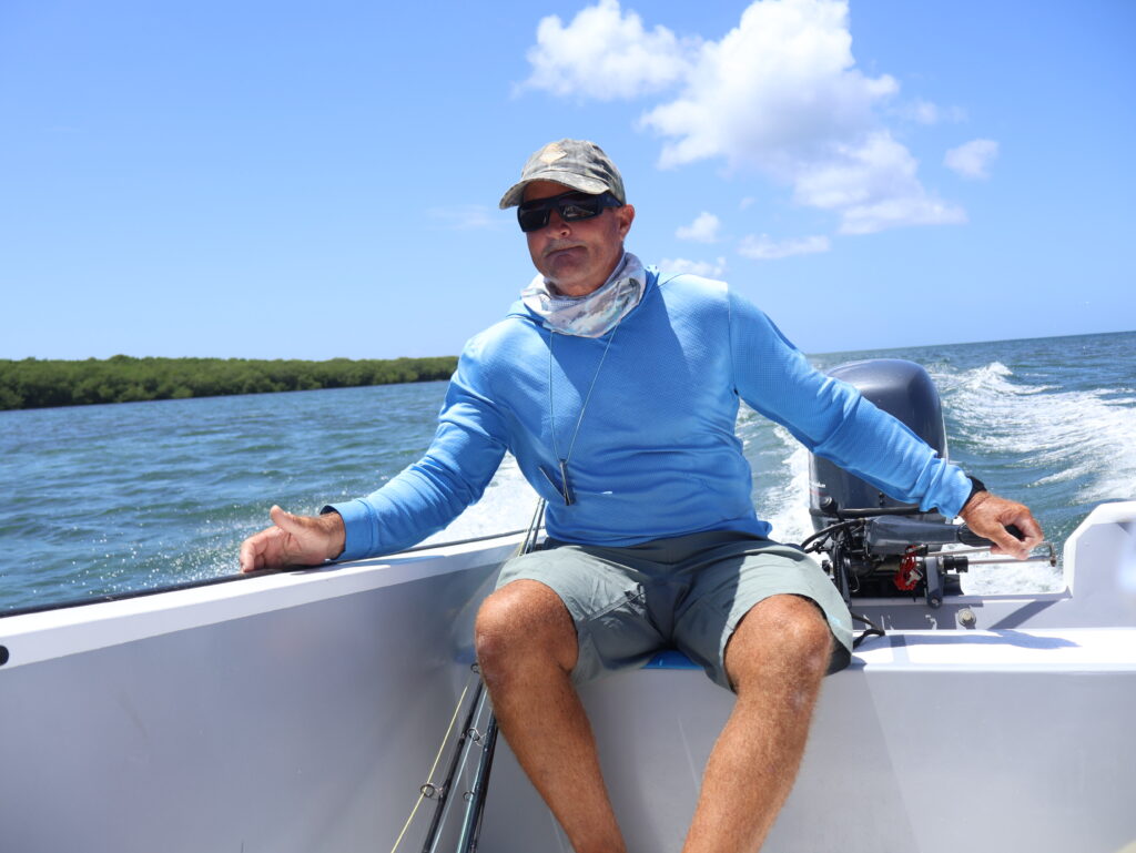 Captain Perry steering the tiller-driven panga along a mangrove shoreline under blue sky